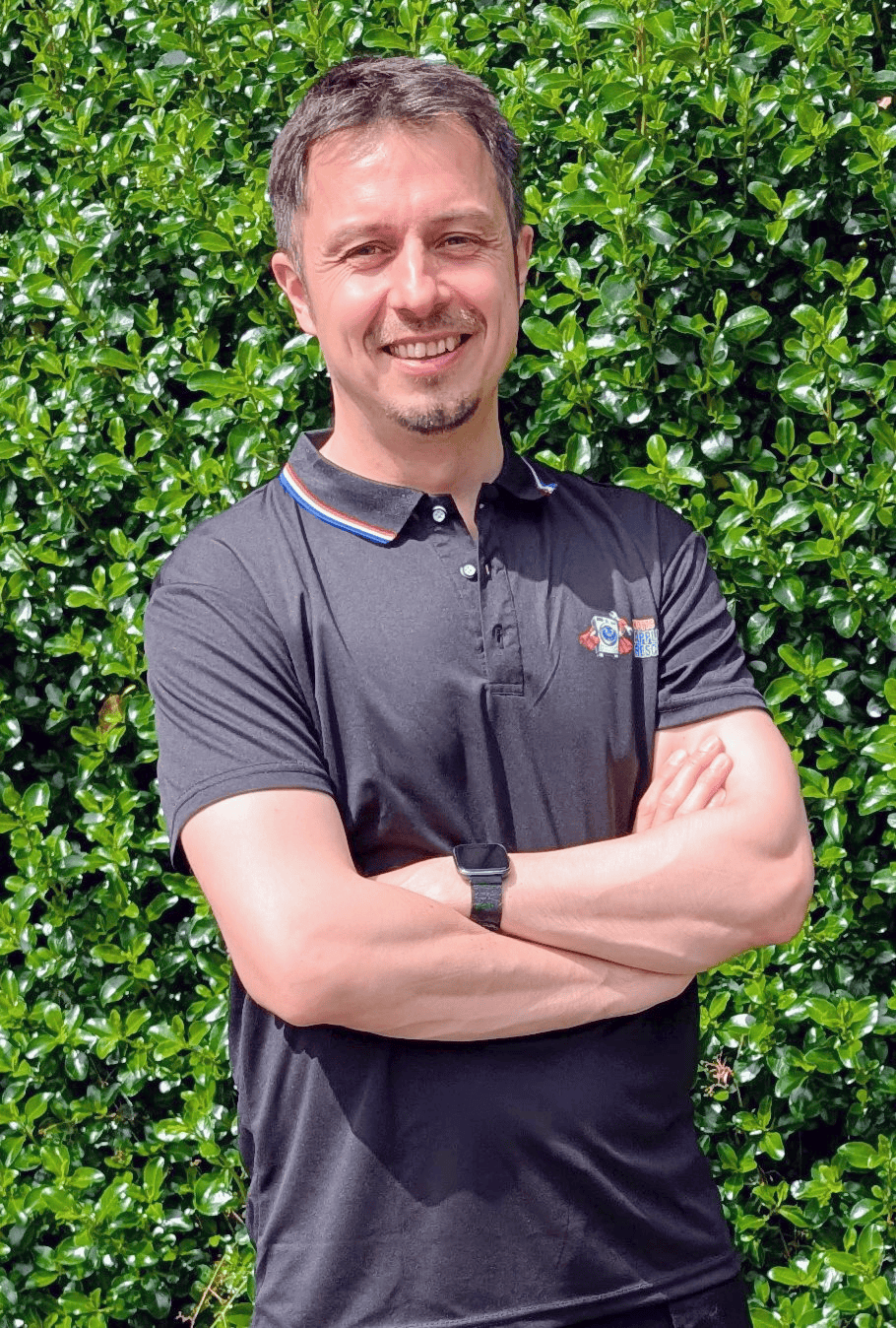 A person stands outside, smiling with arms crossed, wearing a black short-sleeved polo shirt and a smartwatch—ready to assist with appliance repair Nottingham. Green leafy bushes fill the background.